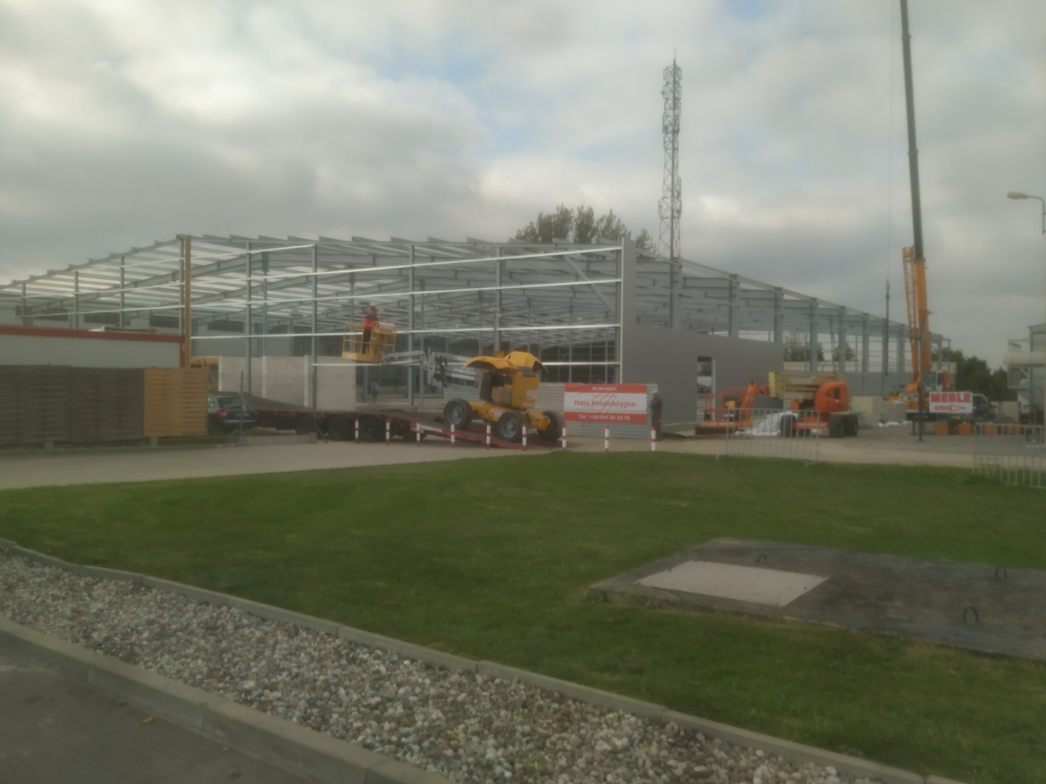 Montaż stalowej konstrukcji hali produkcyjnej, widoczny szkielet budynku, żuraw budowlany i podnośnik koszowy na tle pochmurnego nieba.