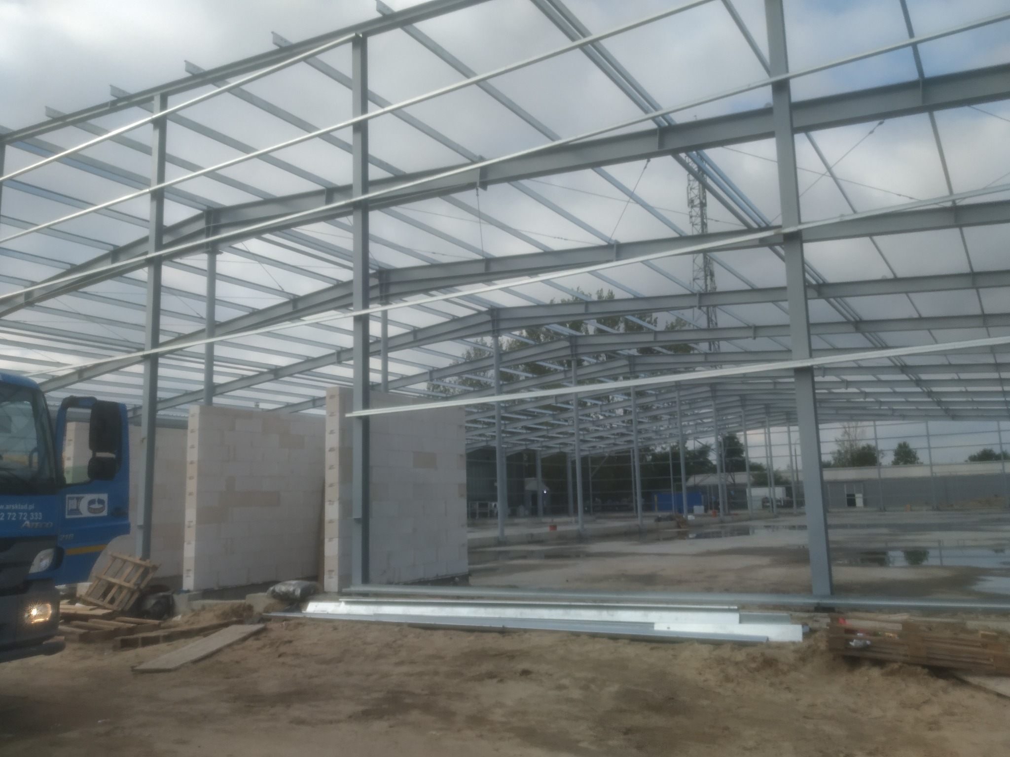 Wznoszenie szkieletu hali z belek stalowych na placu budowy, widoczny częściowo niebieski samochód dostawczy i bloczki betonowe.