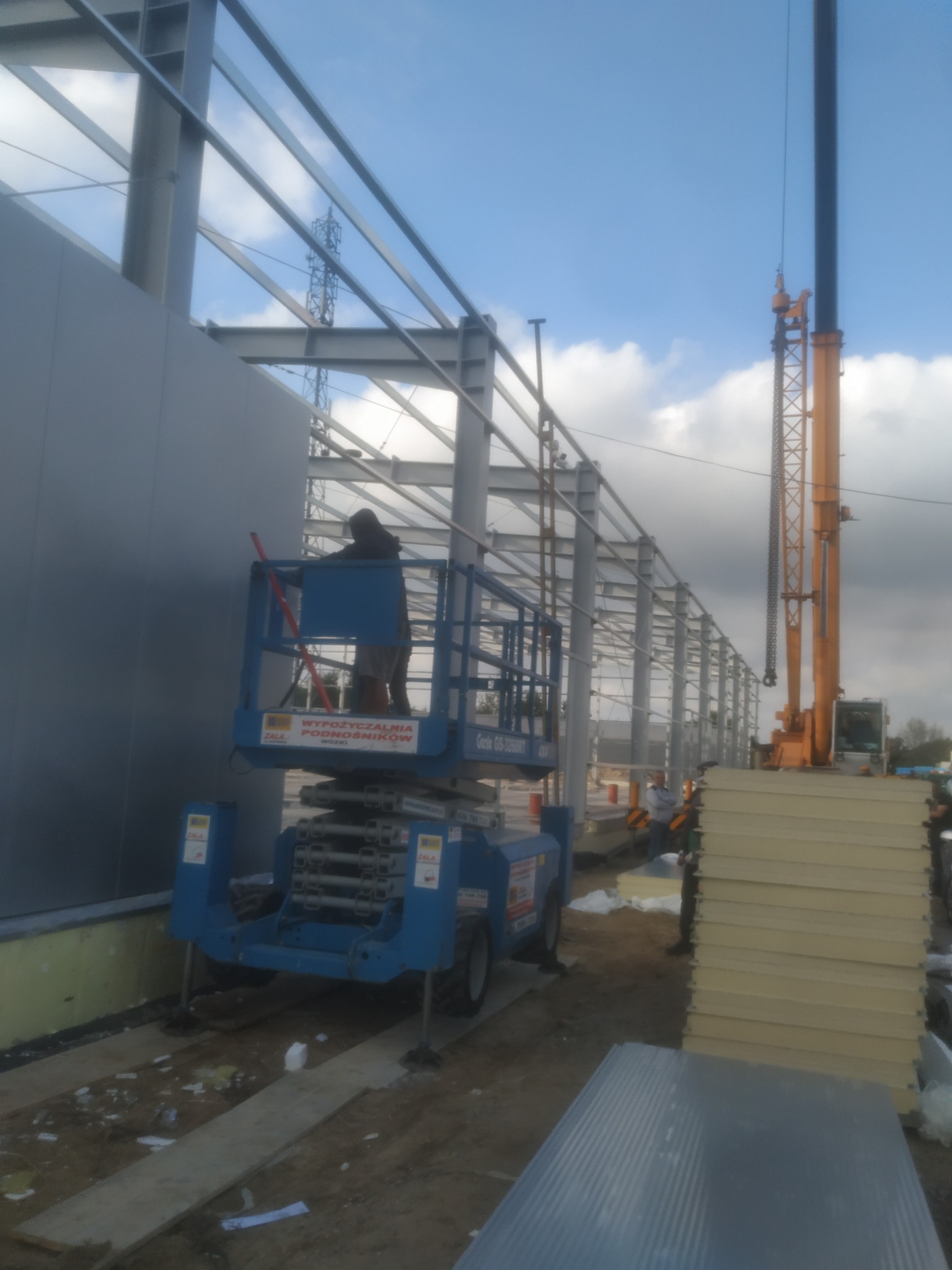 Montaż stalowej konstrukcji hali: pracownik na podnośniku koszowym przytwierdza panele ścienne, w tle widoczny dźwig i elementy konstrukcji szkieletowej budynku.