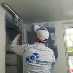 Pracownik w firmowej odzieży nakłada masę szpachlową na ścianę przy użyciu długiej szpachelki, widoczne ślady po wcześniejszym malowaniu na niebiesko.