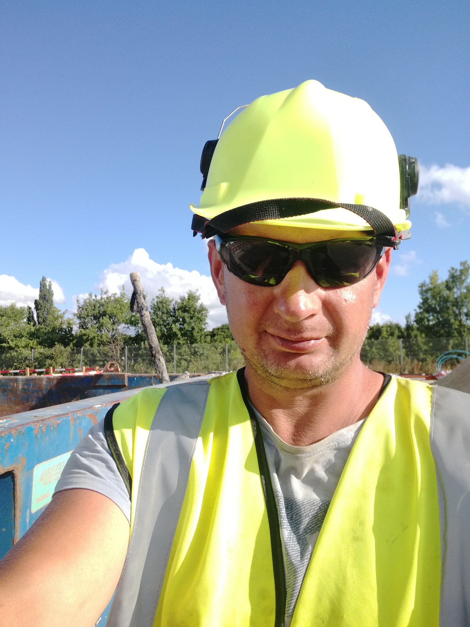 Autoportret pracownika budowlanego w kasku i kamizelce odblaskowej na tle nieba i drzew, z widocznym kontenerem budowlanym.