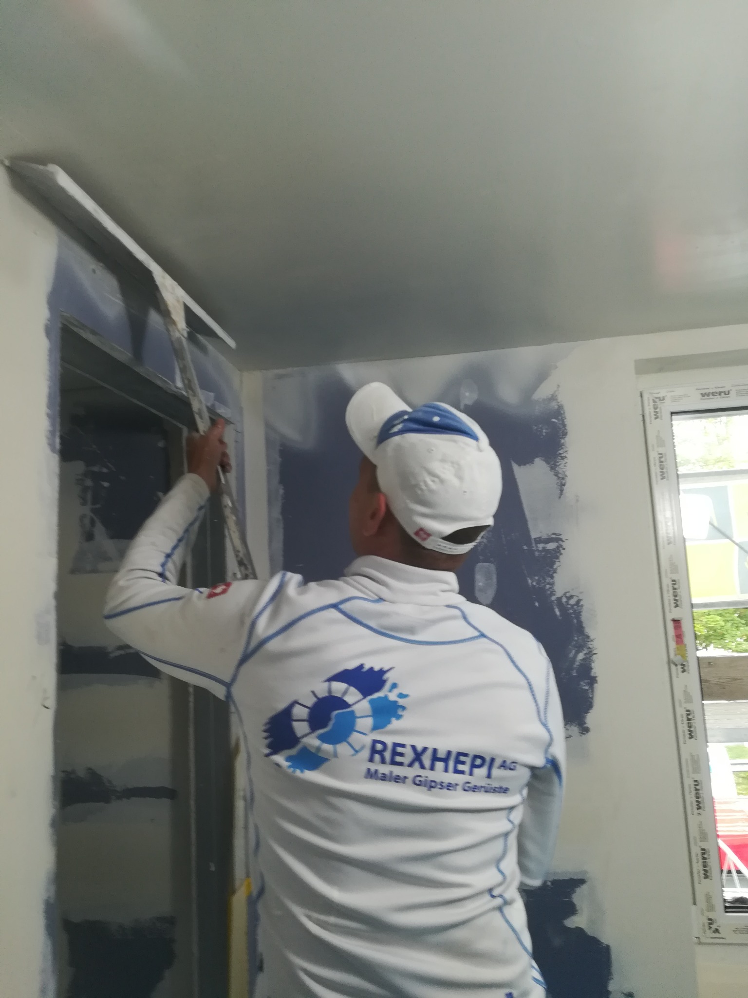 Pracownik w firmowej odzieży nakłada masę szpachlową na ścianę przy użyciu długiej szpachelki, widoczne ślady po wcześniejszym malowaniu na niebiesko.