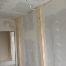 DAR - MAX DARIUSZ NAPIÓRKOWSKI - Ściana z płyt kartonowo-gipsowych w trakcie szpachlowania, widoczne drewniane listwy wzmacniające narożnik, niedokończone wygładzenie powierzchni sufitu i wystający kabel elektryczny.