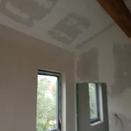 Wnętrze w trakcie prac wykończeniowych: ściany pokryte gładzią gipsową z widocznymi łatami, okno z widokiem na zieleń, drewniany element stropu. Widoczny etap szpachlowania połączeń płyt...