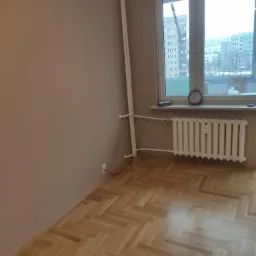 Pomalowane na beżowo wnętrze pokoju z parkietem ułożonym w jodełkę, białym kaloryferem pod oknem i pionowymi rurami grzewczymi w rogu.