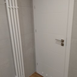 DAR - MAX DARIUSZ NAPIÓRKOWSKI - Biała, pionowa grzejnik drabinkowy na tle szarej ściany z płytek, obok białe drzwi z poziomymi frezami w wykończonej łazience z drewnopodobną podłogą.