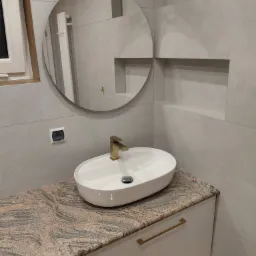 Nowoczesna łazienka z owalną umywalką nablatową, złotą armaturą i okrągłym lustrem z podświetleniem na szarej, gładkiej ścianie.