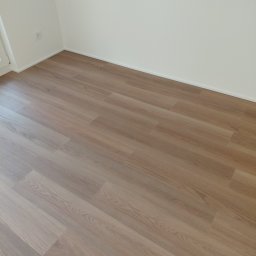 DAR - MAX DARIUSZ NAPIÓRKOWSKI - Jasne panele podłogowe ułożone w nowym pokoju z białymi ścianami i listwami przypodłogowymi. Widoczny fragment gniazdka elektrycznego.