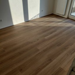 DAR - MAX DARIUSZ NAPIÓRKOWSKI - Nowa, drewnopodobna podłoga w jasnym pokoju z grzejnikiem i oknem. Na ścianie widoczne cienie. Ujęcie z niskiej perspektywy podkreśla wzór paneli.