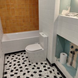 DAR - MAX DARIUSZ NAPIÓRKOWSKI - Biała toaleta w łazience z mozaikową podłogą i pomarańczowymi płytkami nad wanną. Widoczne przyłącza wodne i rolka papieru toaletowego w niszy.