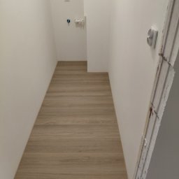 DAR - MAX DARIUSZ NAPIÓRKOWSKI - Wąskie pomieszczenie po remoncie z panelami podłogowymi imitującymi drewno. Białe ściany z instalacją wodną i gniazdkiem elektrycznym. Wykończenie narożnika.
