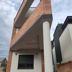 DAR - MAX DARIUSZ NAPIÓRKOWSKI - Dwupiętrowy dom z ceglaną elewacją i kolumną, widok z dołu na balkon z ceglaną balustradą i szarą listwą, w tle błękitne niebo i sąsiedni budynek z szarą fasadą.