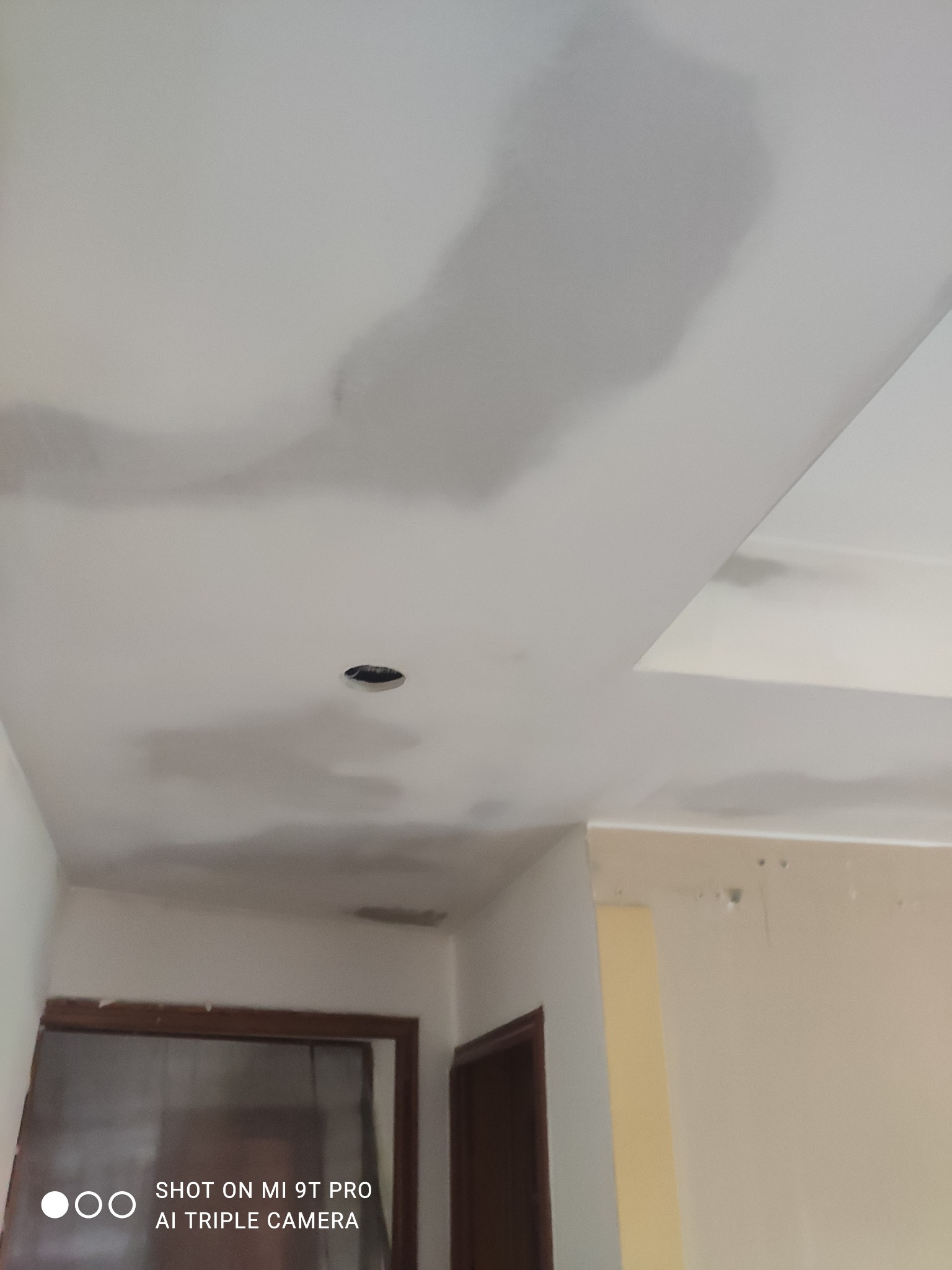 Niedokończone gładzie na suficie z widocznym otworem na oświetlenie, szare plamy na białym tle, widoczne fragmenty ścian i drzwi w tle.