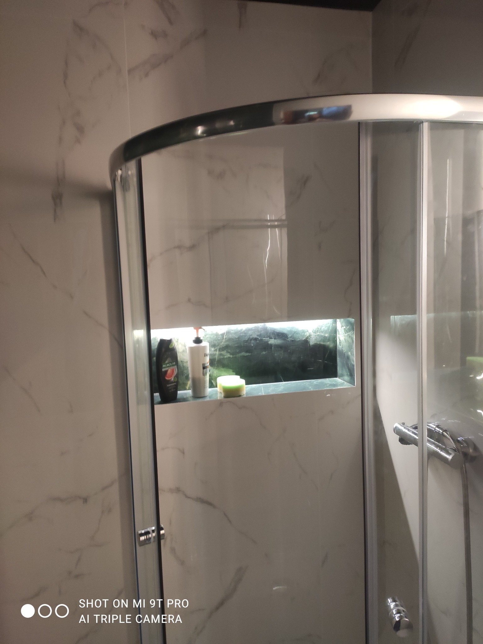 Nowoczesna kabina prysznicowa z półką w ścianie, na której stoją kosmetyki i gąbka; ściany wyłożone płytkami imitującymi marmur.