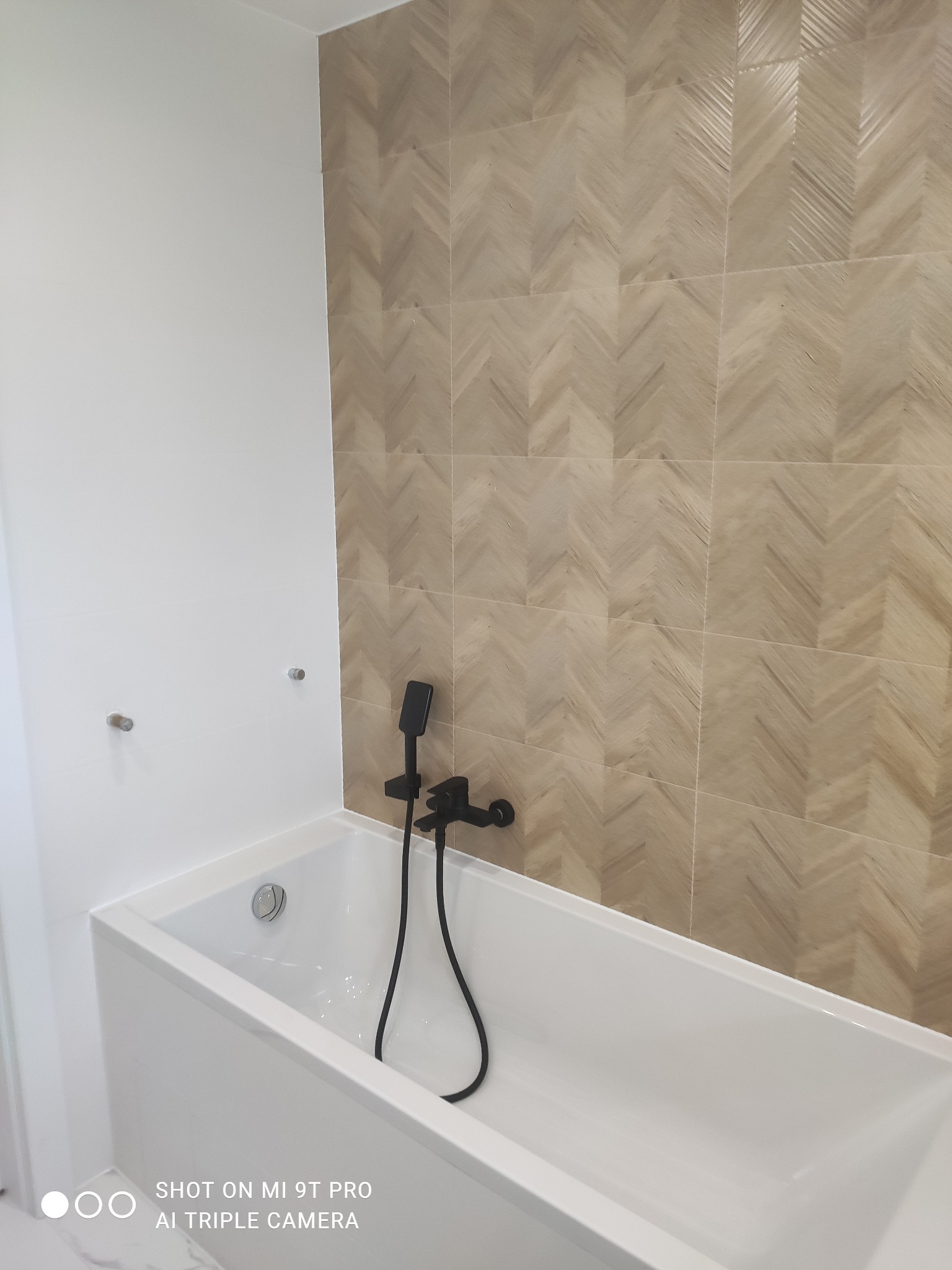 Nowoczesna, biała wanna z czarną armaturą i dekoracyjną ścianą z płytek ułożonych w jodełkę.