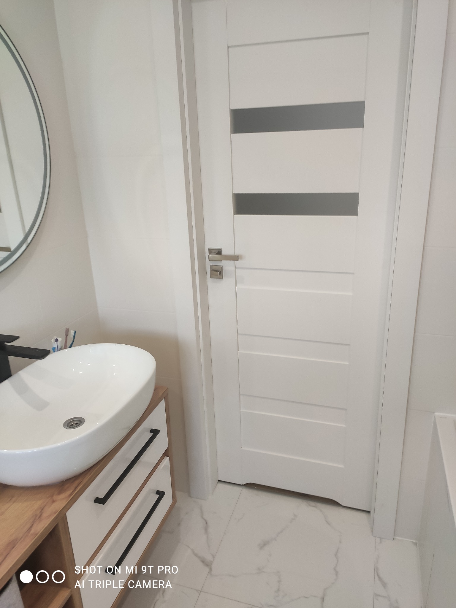 Nowoczesna łazienka z owalną umywalką nablatową, szafką z drewnianym blatem i czarnymi uchwytami, białą ścianą z lustrem w okrągłej ramie oraz białą, panelową ścianą i drzwiami z szarymi szybami.