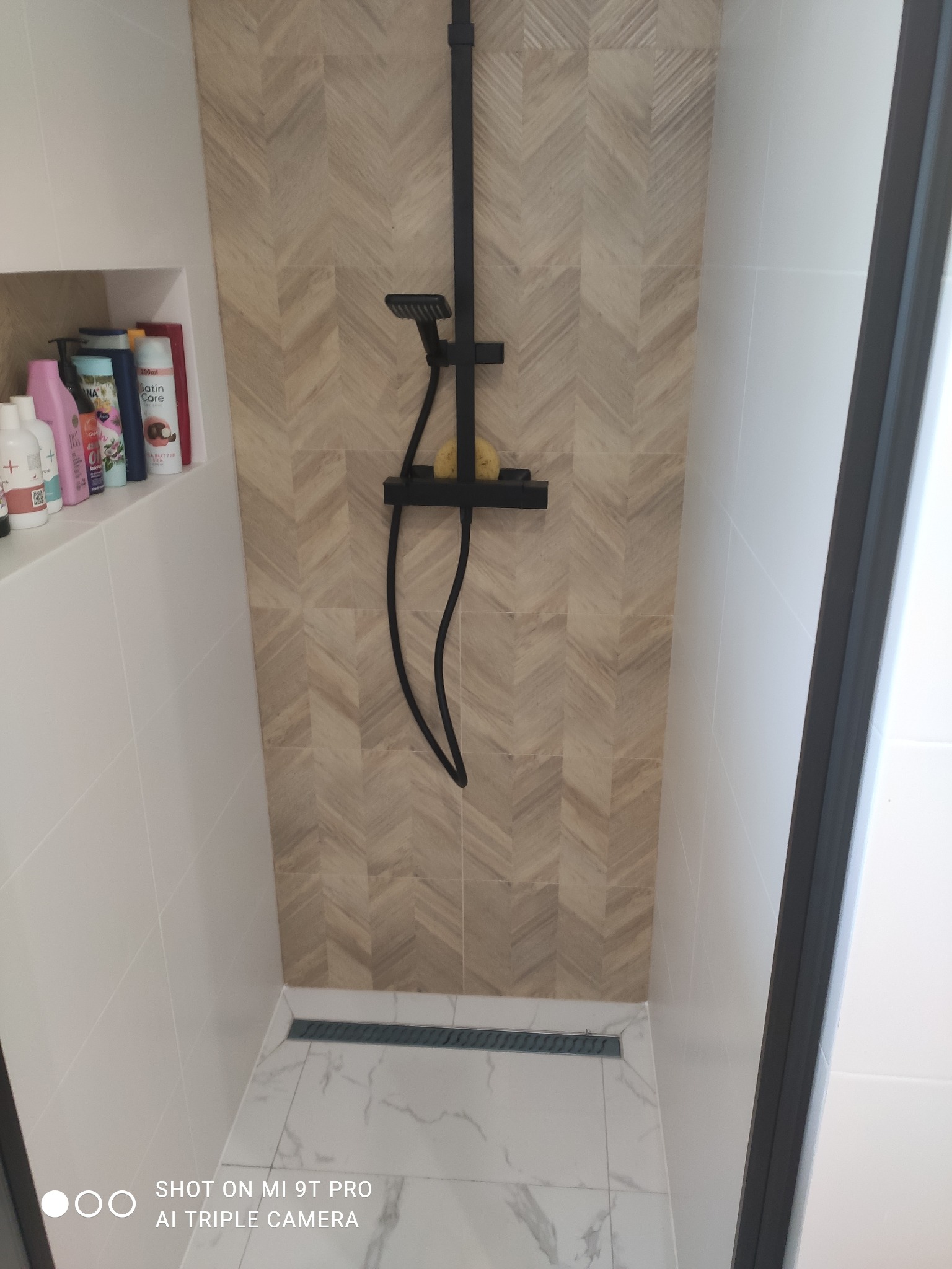 Nowoczesna kabina prysznicowa z czarną armaturą, wykończona płytkami w jodełkę na ścianie i marmurowymi płytkami na podłodze z odpływem liniowym.