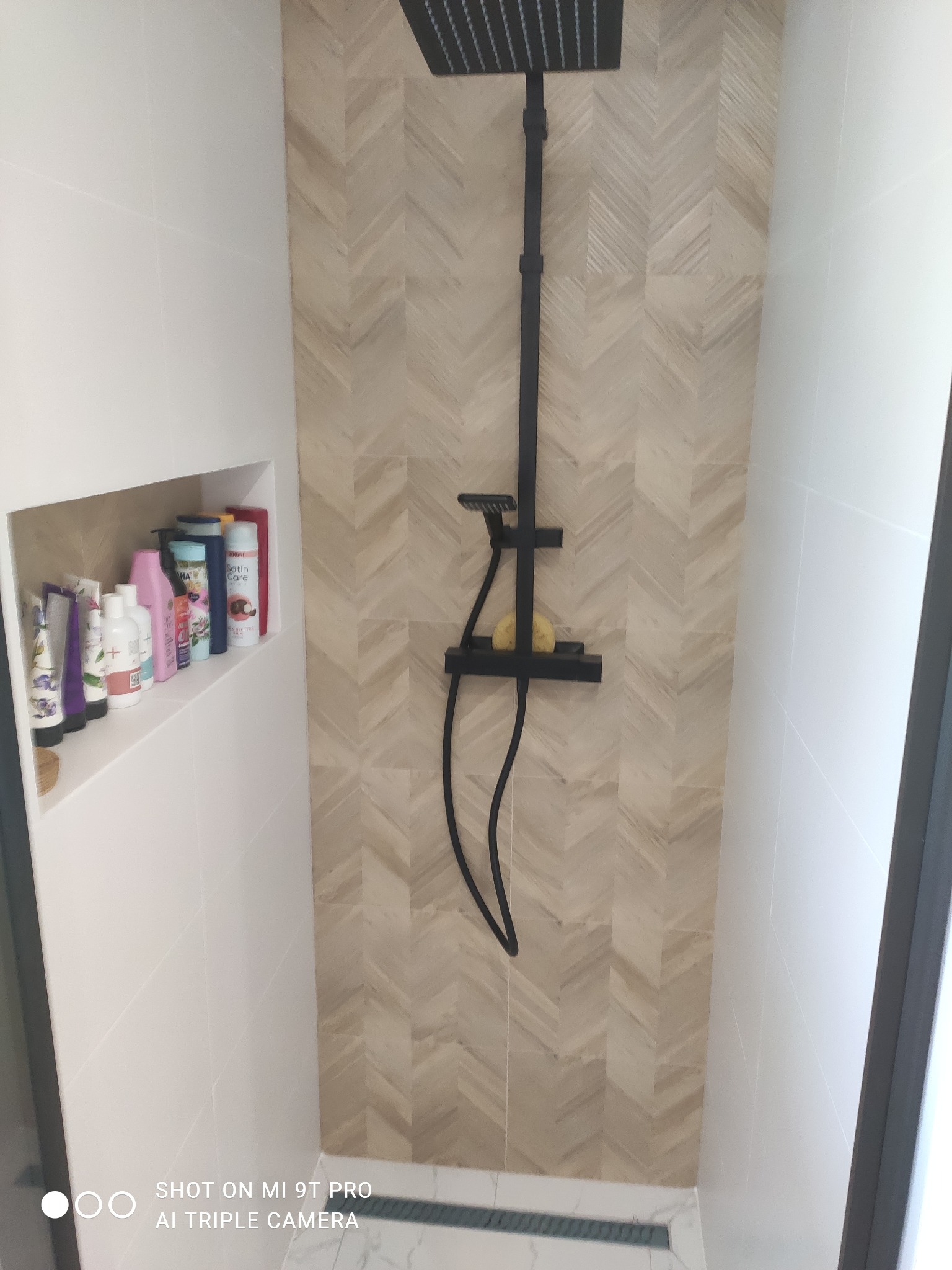 Kabina prysznicowa z czarną armaturą, wykończona płytkami ułożonymi w jodełkę, z wnęką na kosmetyki po lewej stronie.