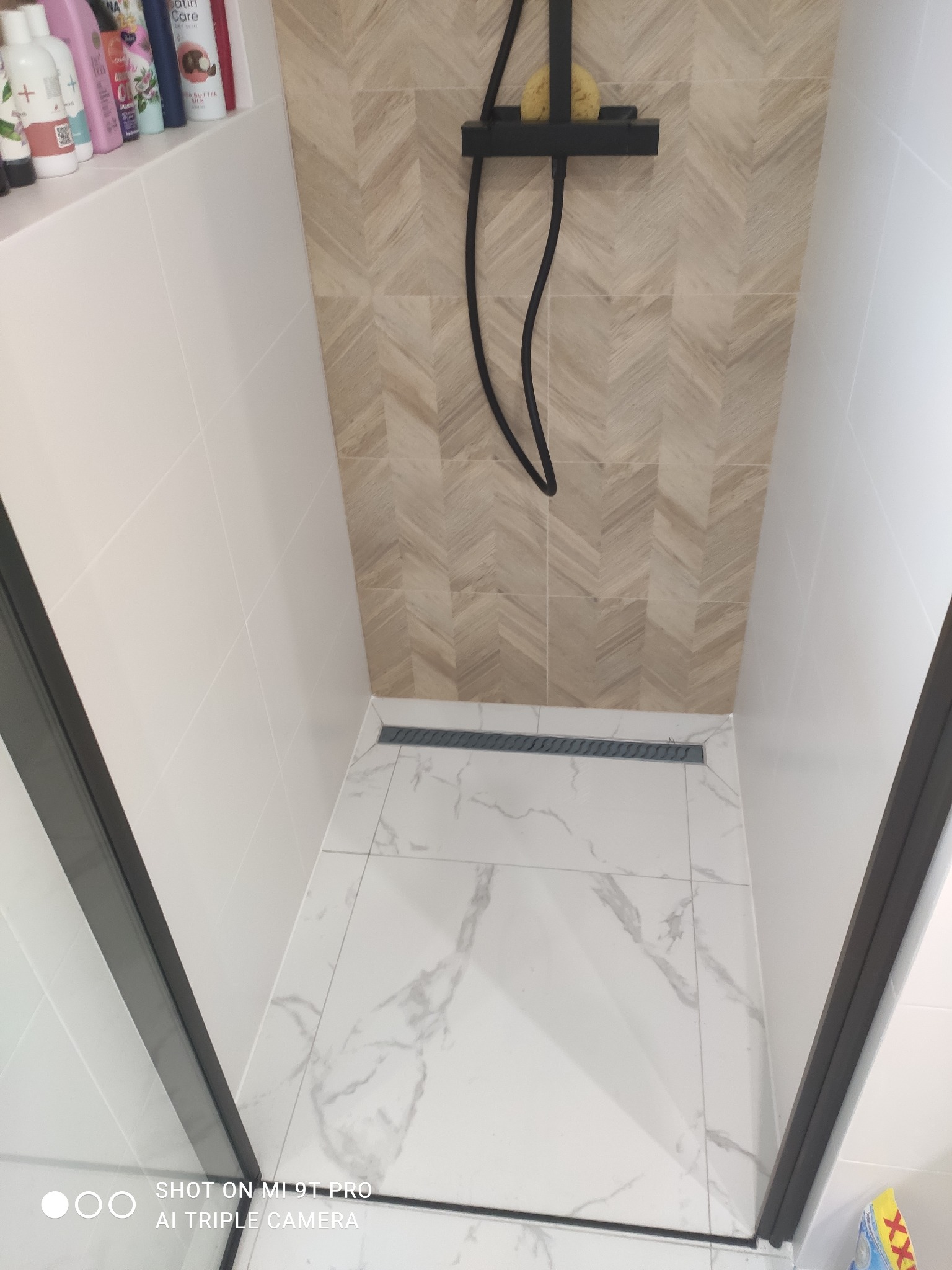 Nowoczesny prysznic walk-in z odpływem liniowym, wykończony płytkami imitującymi drewno na ścianie i białym marmurem na podłodze, czarna armatura i szklana ściana kabiny.