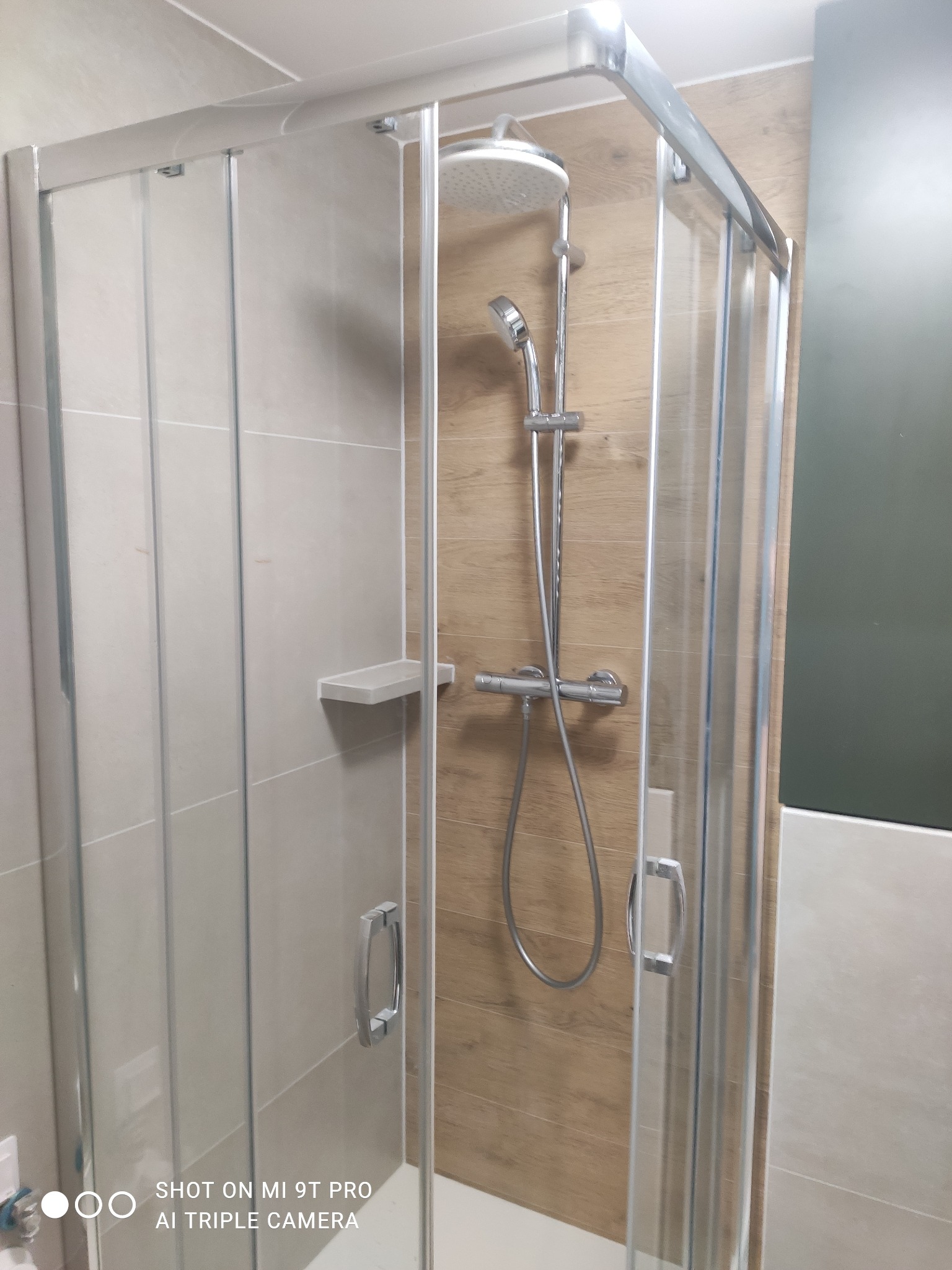 Nowoczesna kabina prysznicowa z przesuwnymi drzwiami, wykończona płytkami imitującymi drewno i jasnobeżowymi płytkami na ścianach, z chromowaną armaturą i małą półką na kosmetyki.