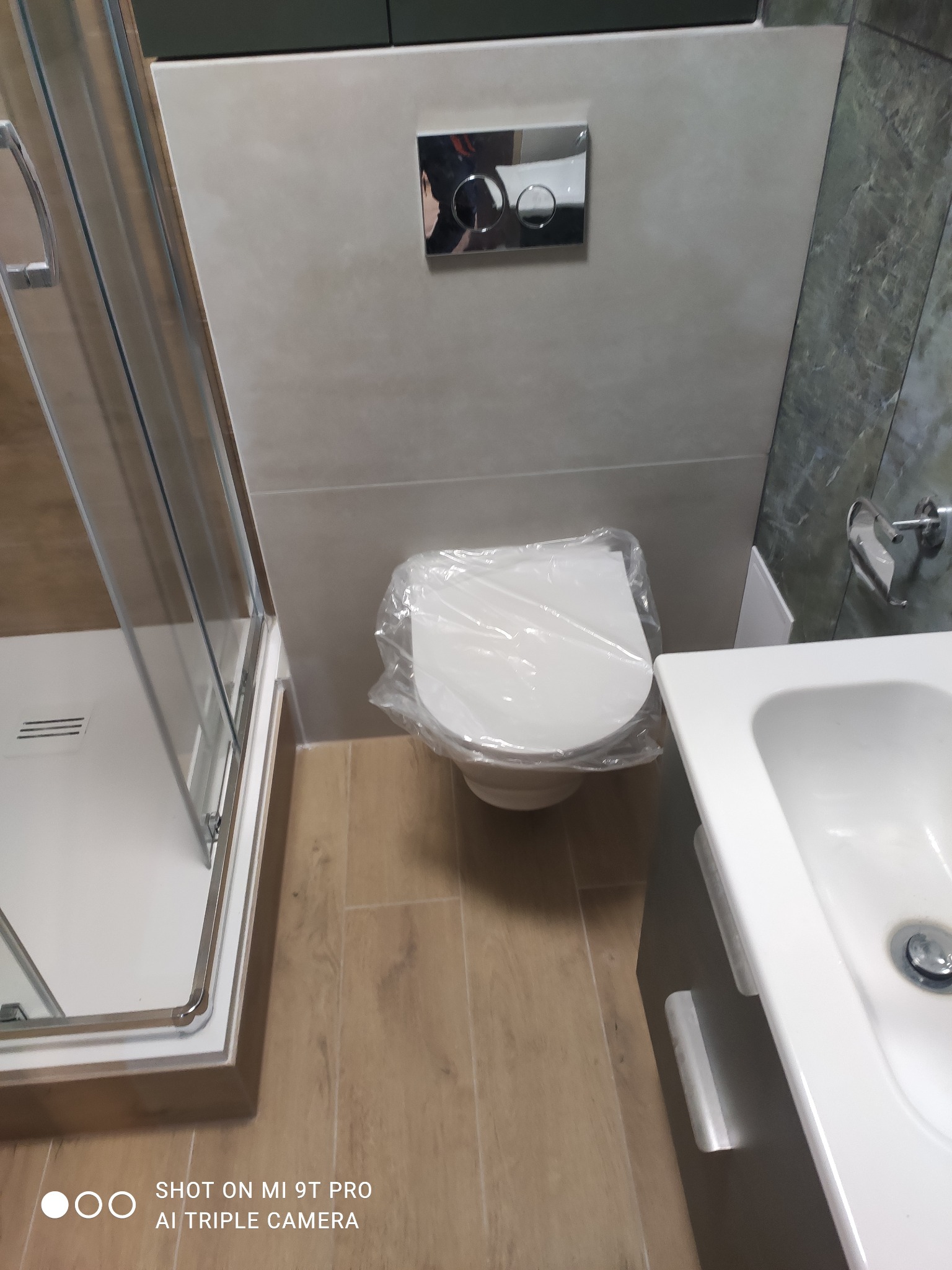 Nowa, biała toaleta zabezpieczona folią ochronną, umywalka i kabina prysznicowa w wykończonej łazience z drewnopodobną podłogą i szarą mozaiką.