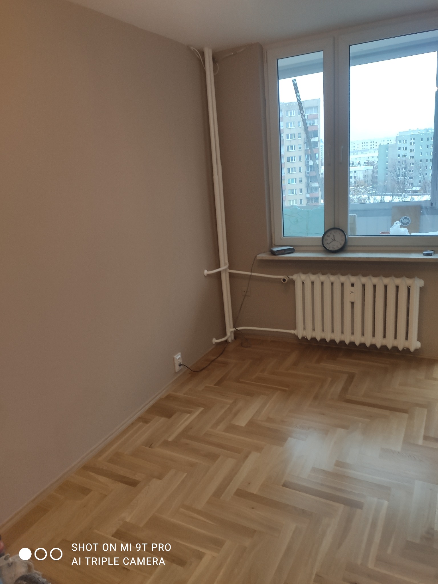 Pomalowane na beżowo wnętrze pokoju z parkietem ułożonym w jodełkę, białym kaloryferem pod oknem i pionowymi rurami grzewczymi w rogu.