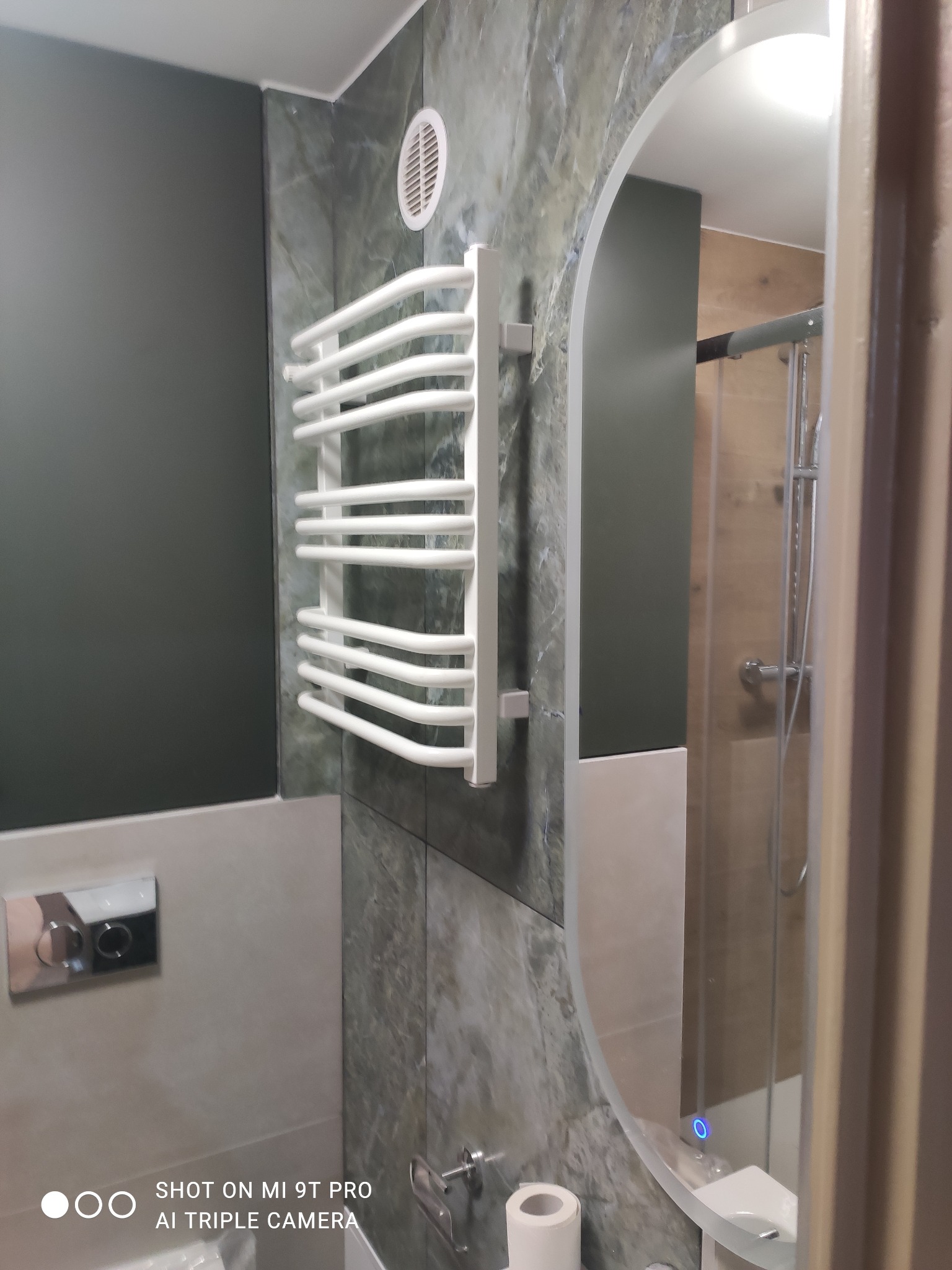 Nowoczesna łazienka z grzejnikiem drabinkowym na ścianie z efektem marmuru, okrągłe lustro z podświetleniem LED i widoczną kabiną prysznicową.