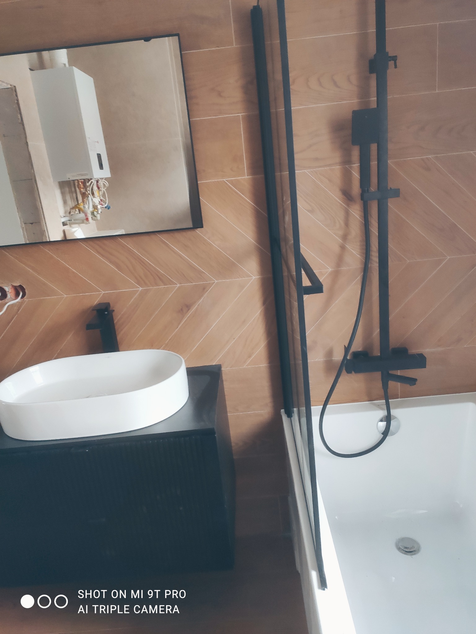 Ukończona łazienka z brązowymi płytkami ułożonymi w jodełkę, czarną armaturą, białą umywalką nablatową i lustrem odbijającym bojler.