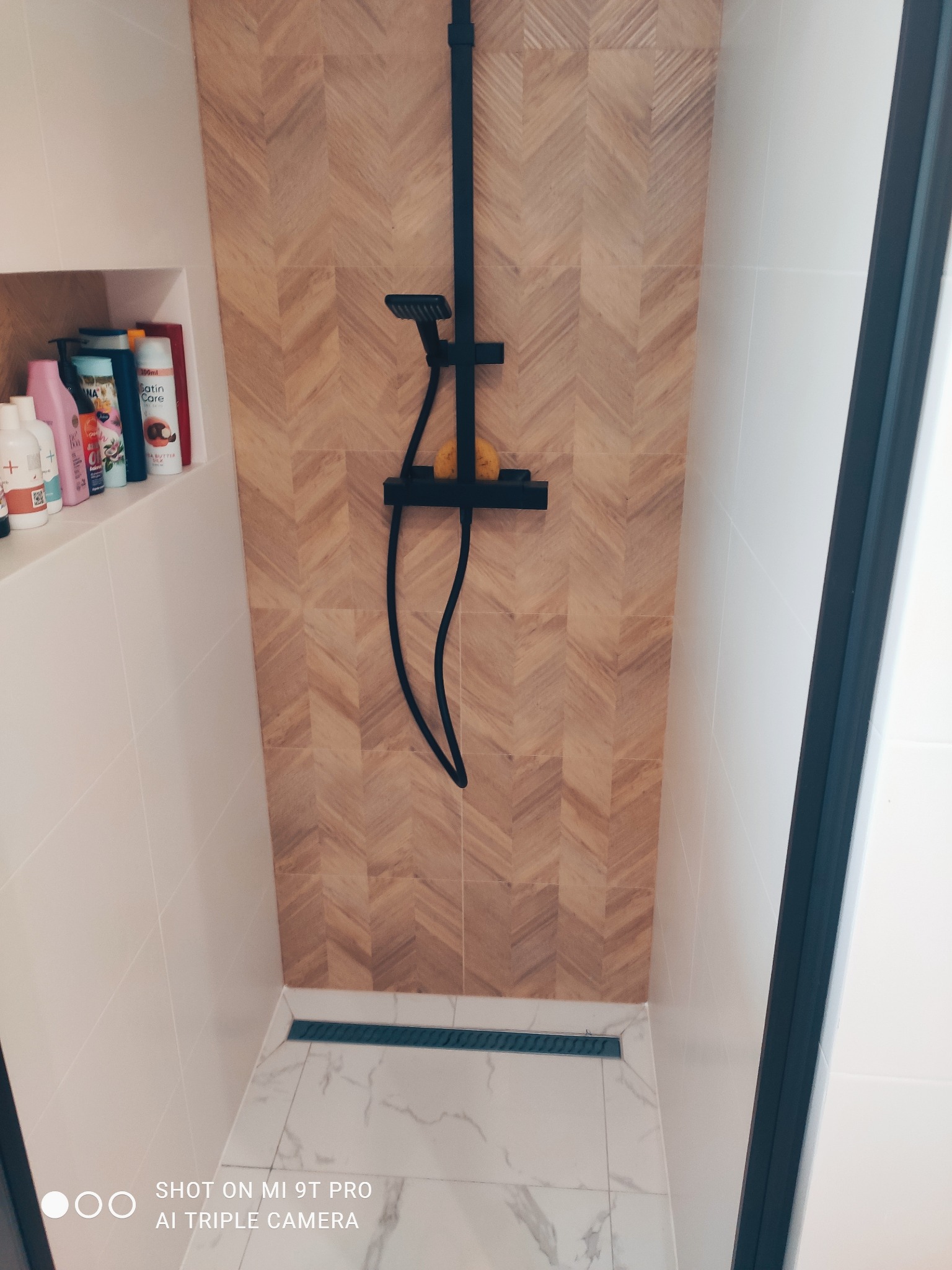 Nowoczesna kabina prysznicowa z drewnopodobną ścianą w jodełkę, czarną armaturą i odpływem liniowym na marmurowej podłodze; wnęka z kosmetykami po lewej stronie.