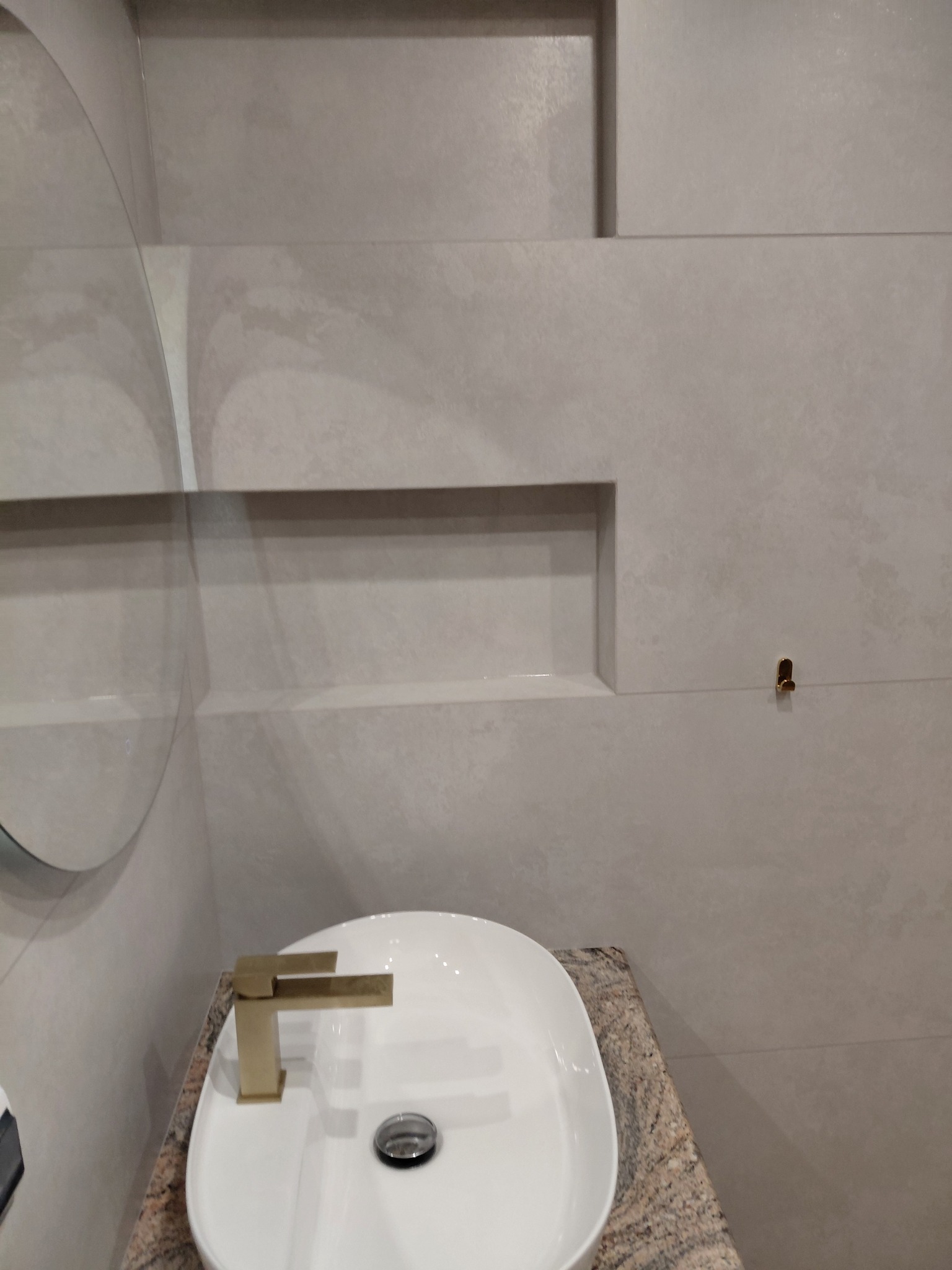 Nowoczesna łazienka: owalna umywalka na blacie z granitu, bateria w kolorze złota, duże szare płytki z wnękami i okrągłe lustro. Minimalistyczny design.