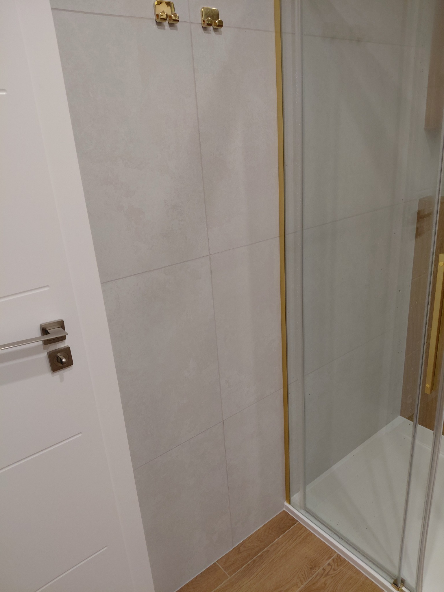 Wykończona kabina prysznicowa z złotymi elementami, jasne płytki na ścianach i drewnopodobna podłoga. Widoczny fragment białych drzwi z metalową klamką.