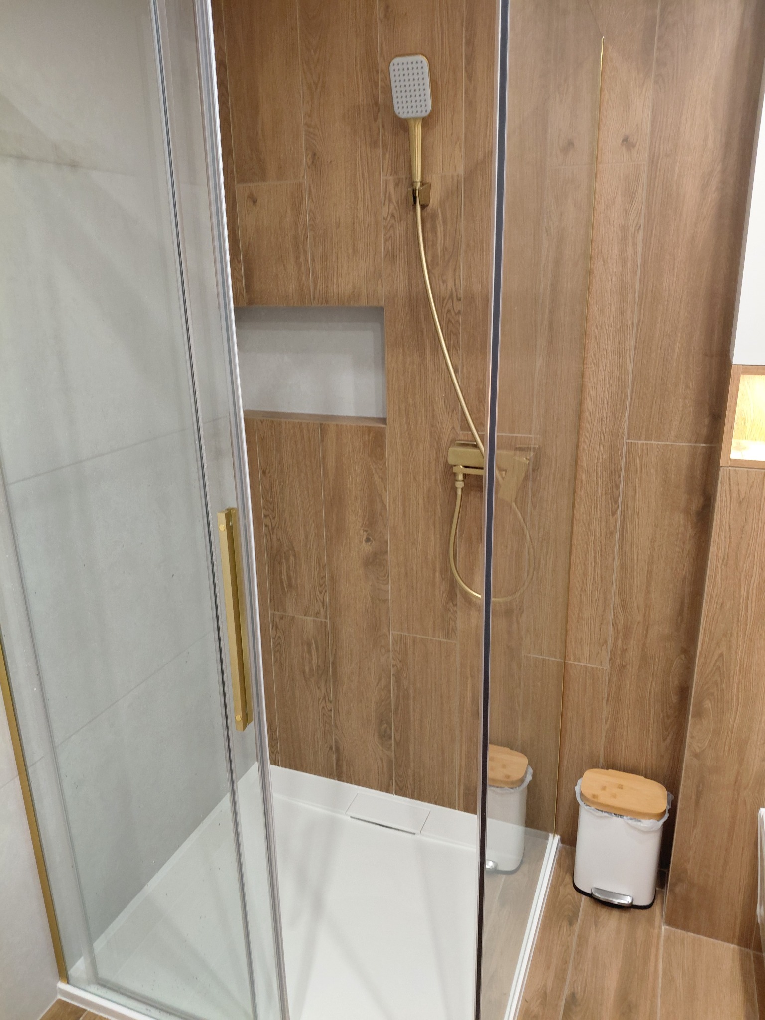 Kabina prysznicowa z deszczownicą w złotej tonacji, drewnopodobne płytki na ścianach, akcesoria łazienkowe w minimalistycznym stylu. Nowoczesne wykończenie łazienki.