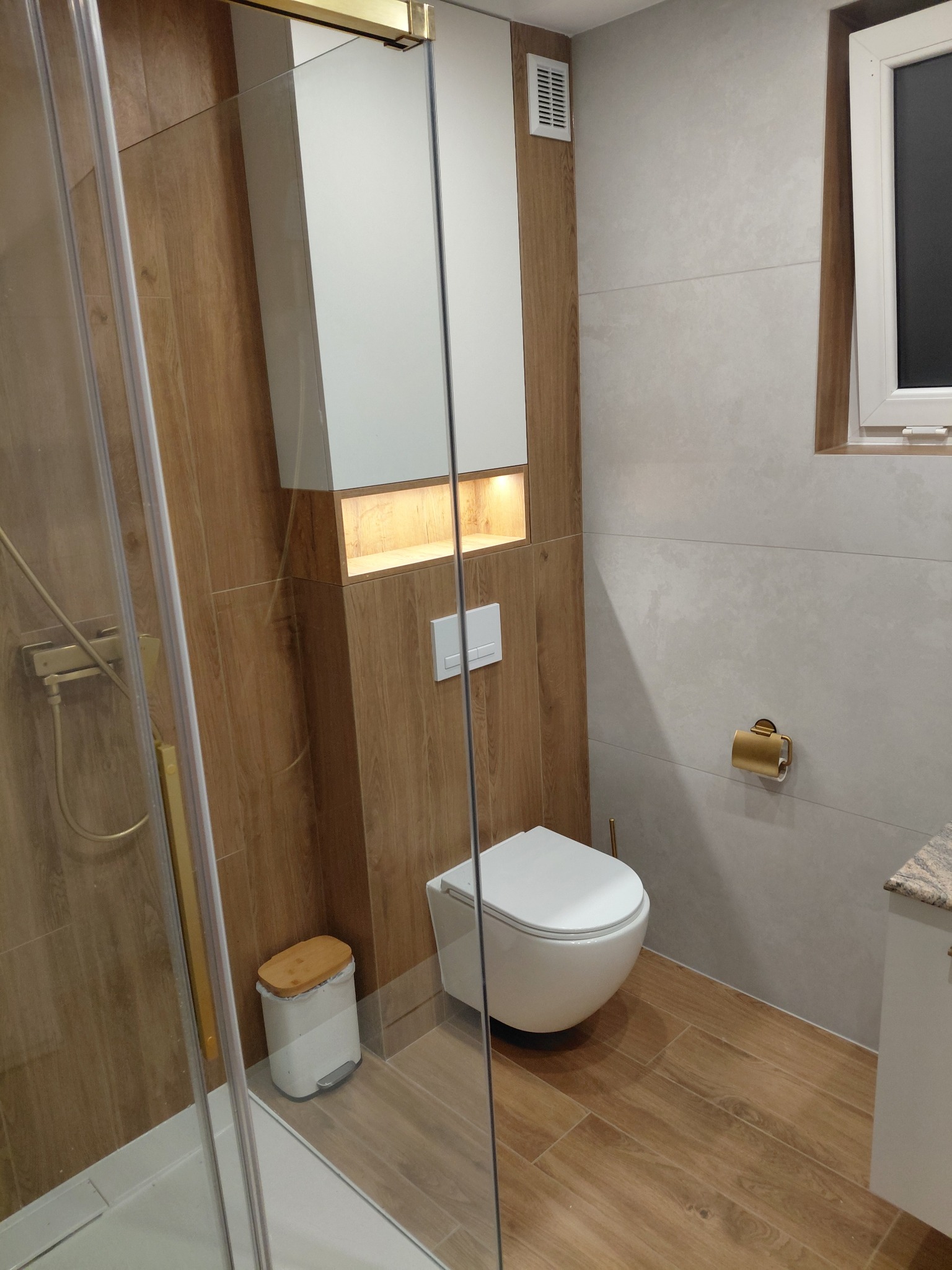 Nowoczesna łazienka z kabiną prysznicową, toaletą i szafką w zabudowie. Płytki imitujące drewno na podłodze i ścianie. Złote akcenty armatury.