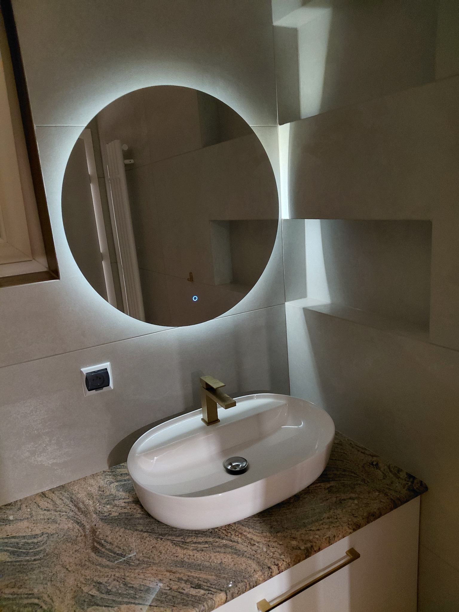 Nowoczesna łazienka: owalne umywalka nablatowa z złotą armaturą na blacie z granitu, okrągłe lustro z podświetleniem LED i geometryczne płytki na ścianie.