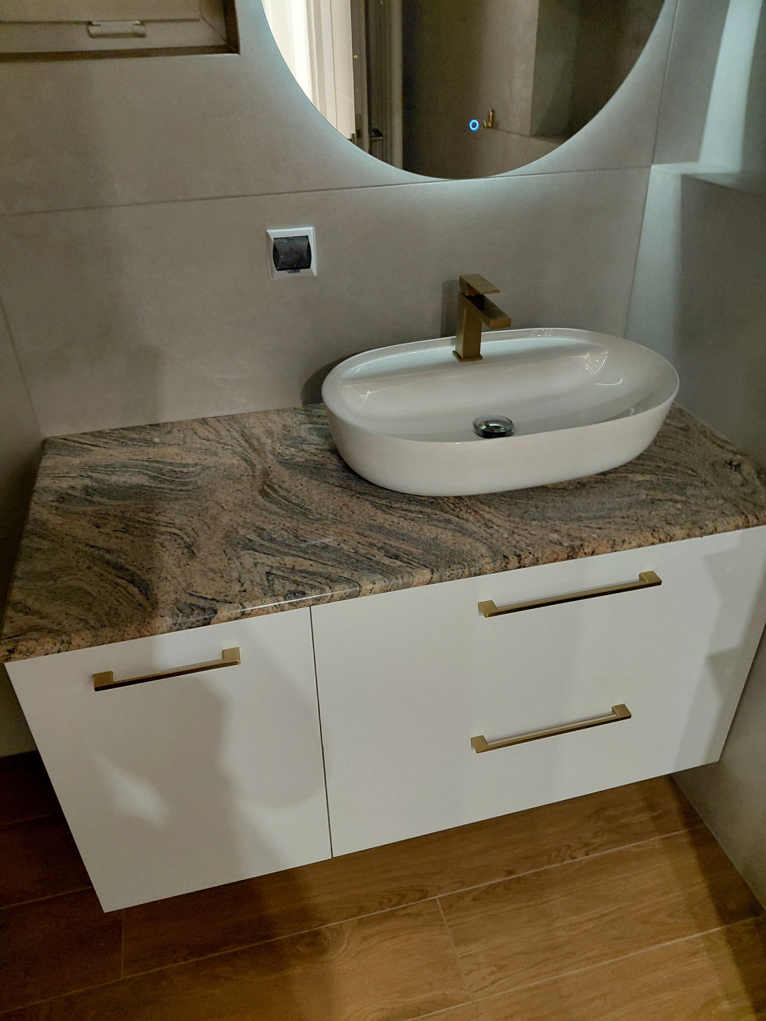 Nowoczesna łazienka: biała umywalka nablatowa z złotą armaturą na szafce z blatem imitującym marmur, okrągłe lustro z podświetleniem LED.