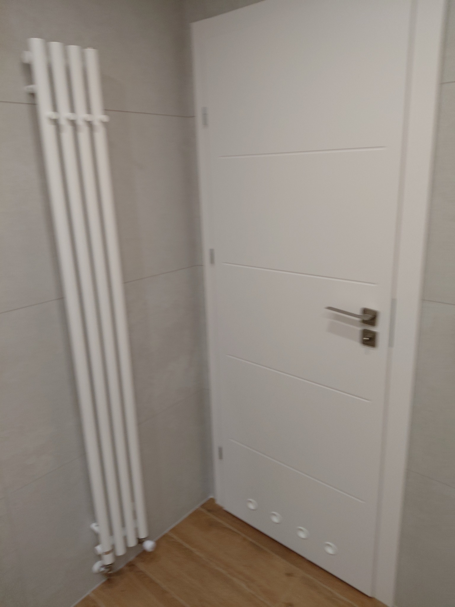 Biała, pionowa grzejnik drabinkowy na tle szarej ściany z płytek, obok białe drzwi z poziomymi frezami w wykończonej łazience z drewnopodobną podłogą.