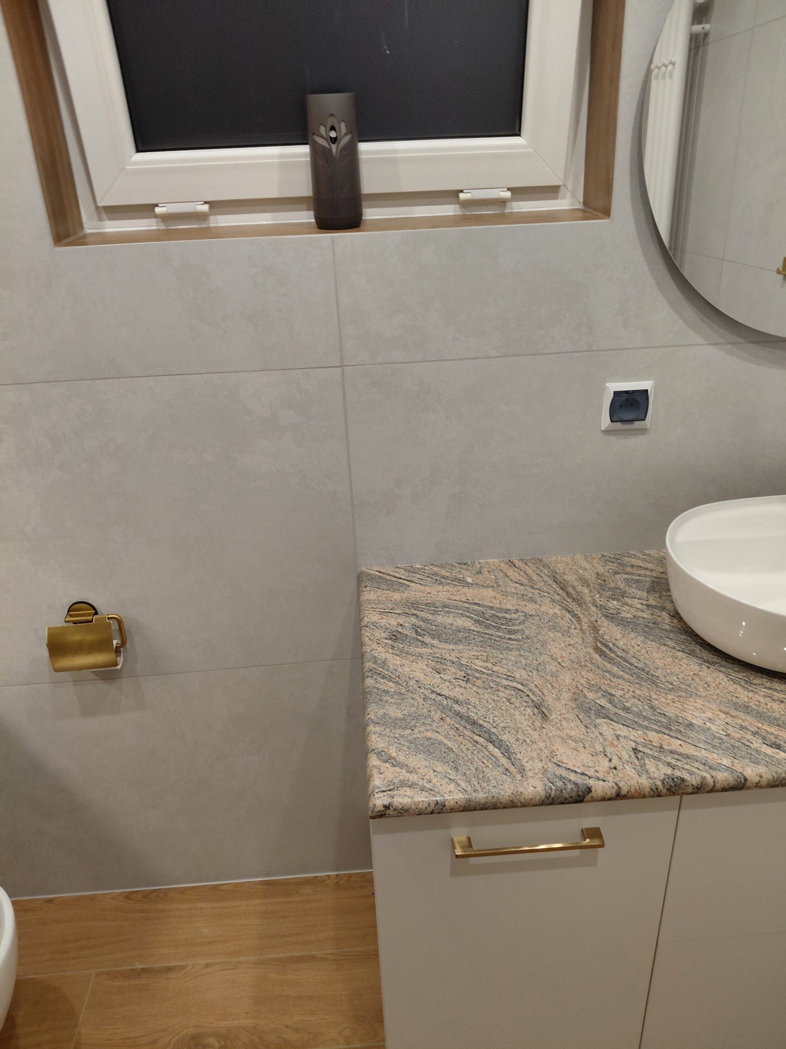 Wykończona łazienka z beżowymi płytkami, drewnopodobną podłogą i blatem z granitu. Widoczny fragment okrągłego lustra i dozownik zapachu na parapecie.