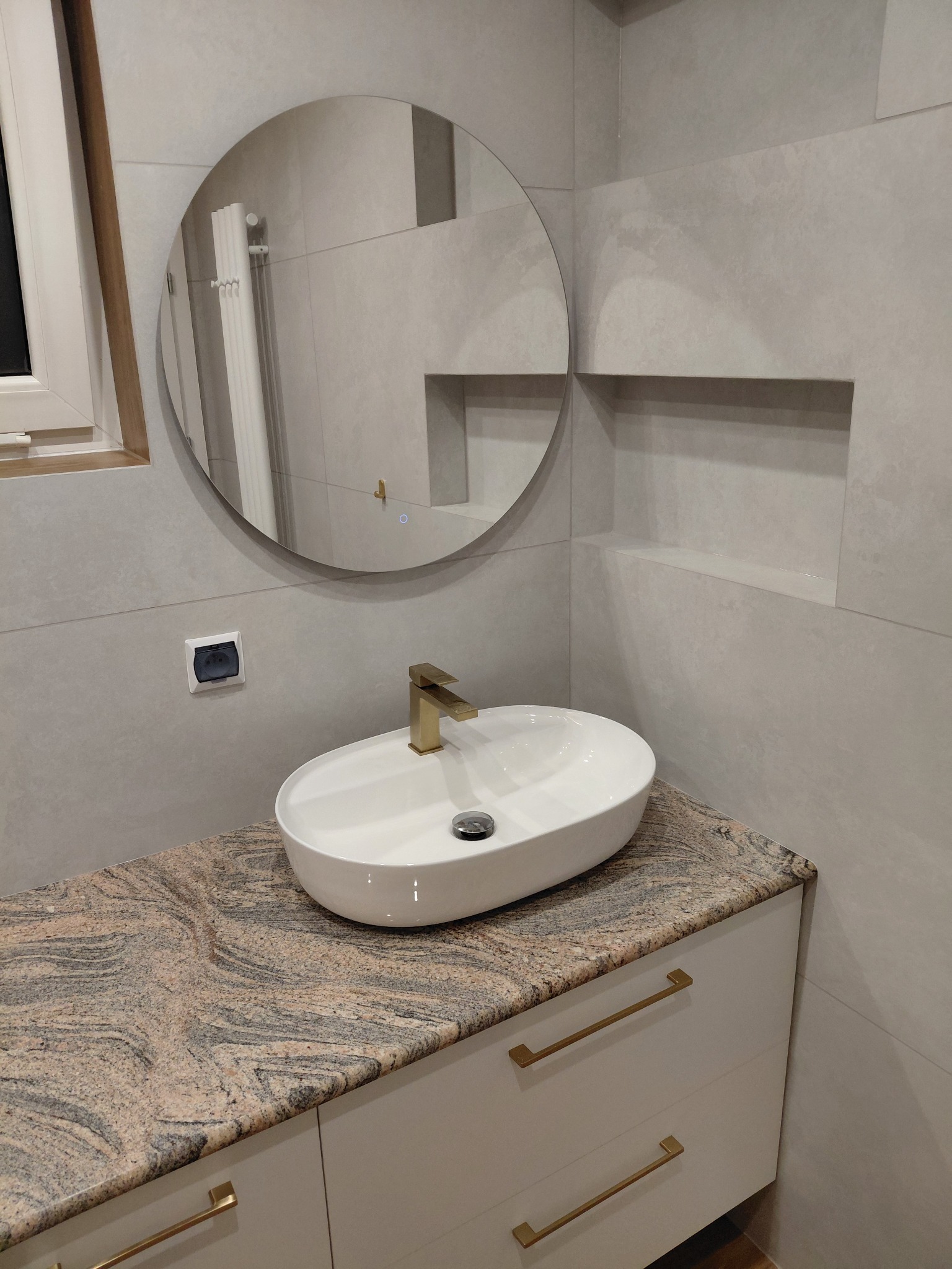 Nowoczesna łazienka z owalną umywalką nablatową, złotą armaturą i okrągłym lustrem z podświetleniem na szarej, gładkiej ścianie.