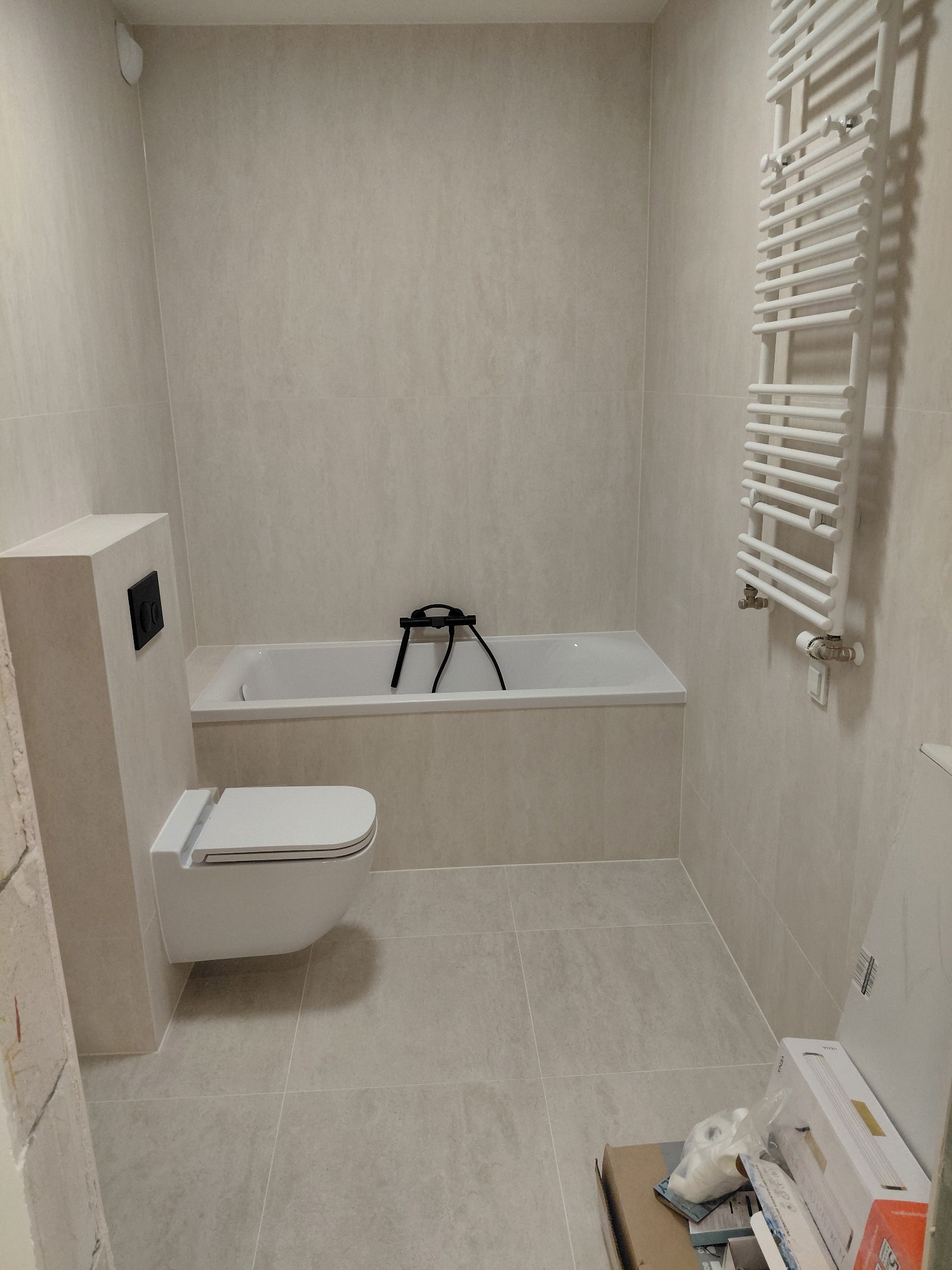 Jasna łazienka po remoncie: beżowe płytki, podwieszana toaleta z czarnym panelem, biała drabinka grzejnikowa i wanna. Widoczne kartony po materiałach.
