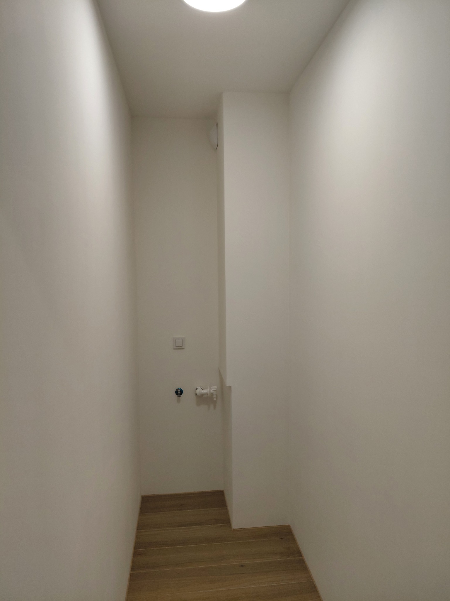 Wnętrze małej, wąskiej łazienki po remoncie. Widoczne białe ściany, drewnopodobna podłoga, włącznik światła i przyłącze wodne. Minimalistyczny styl.