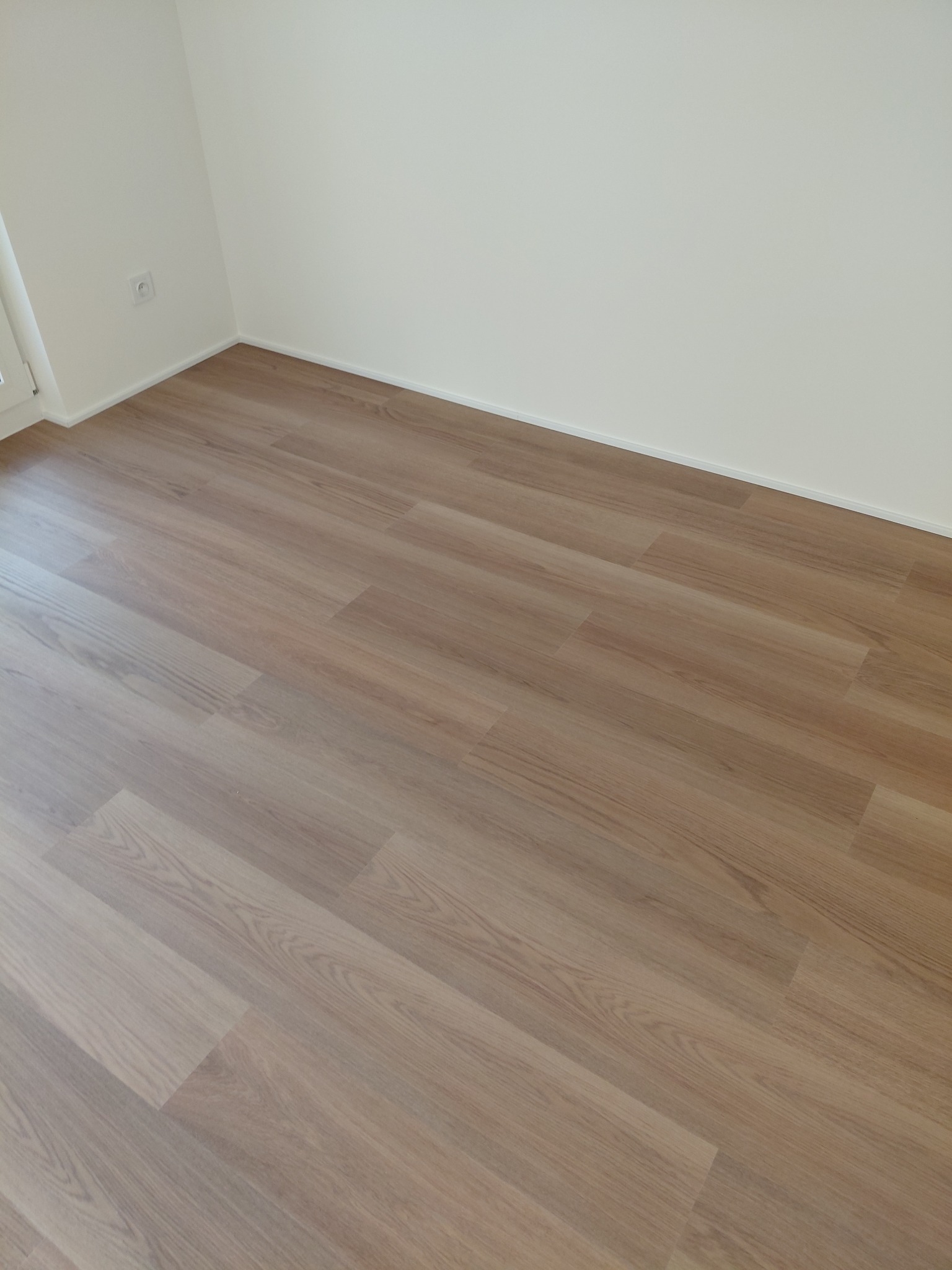 Jasne panele podłogowe ułożone w nowym pokoju z białymi ścianami i listwami przypodłogowymi. Widoczny fragment gniazdka elektrycznego.