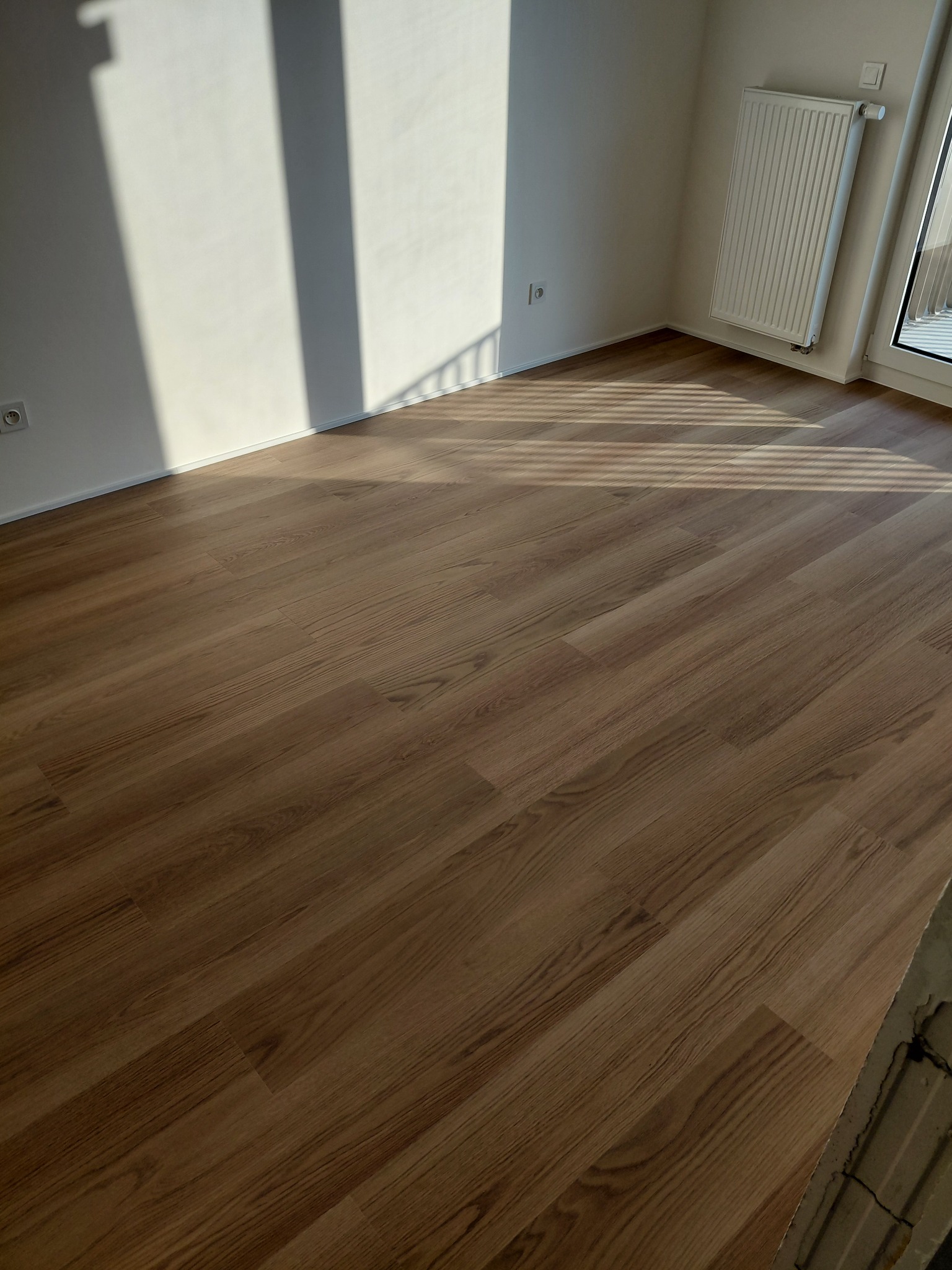 Nowa, drewnopodobna podłoga w jasnym pokoju z grzejnikiem i oknem. Na ścianie widoczne cienie. Ujęcie z niskiej perspektywy podkreśla wzór paneli.