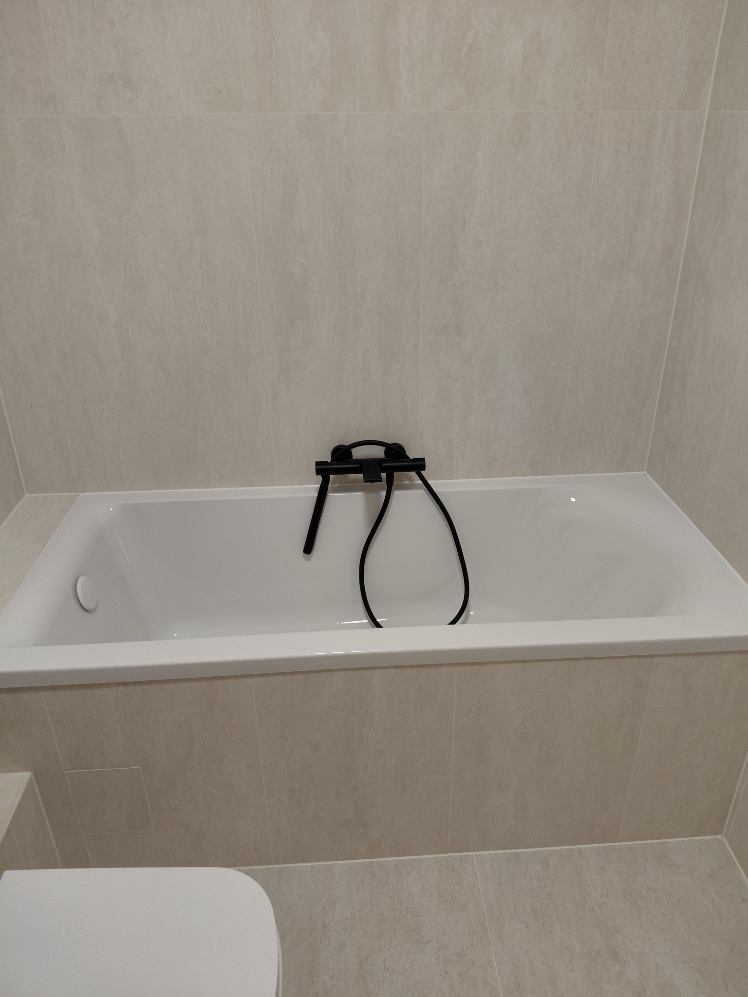 Biała wanna z czarną armaturą na tle beżowych płytek w łazience. Minimalistyczny design, stonowana kolorystyka, widoczny fragment toalety.