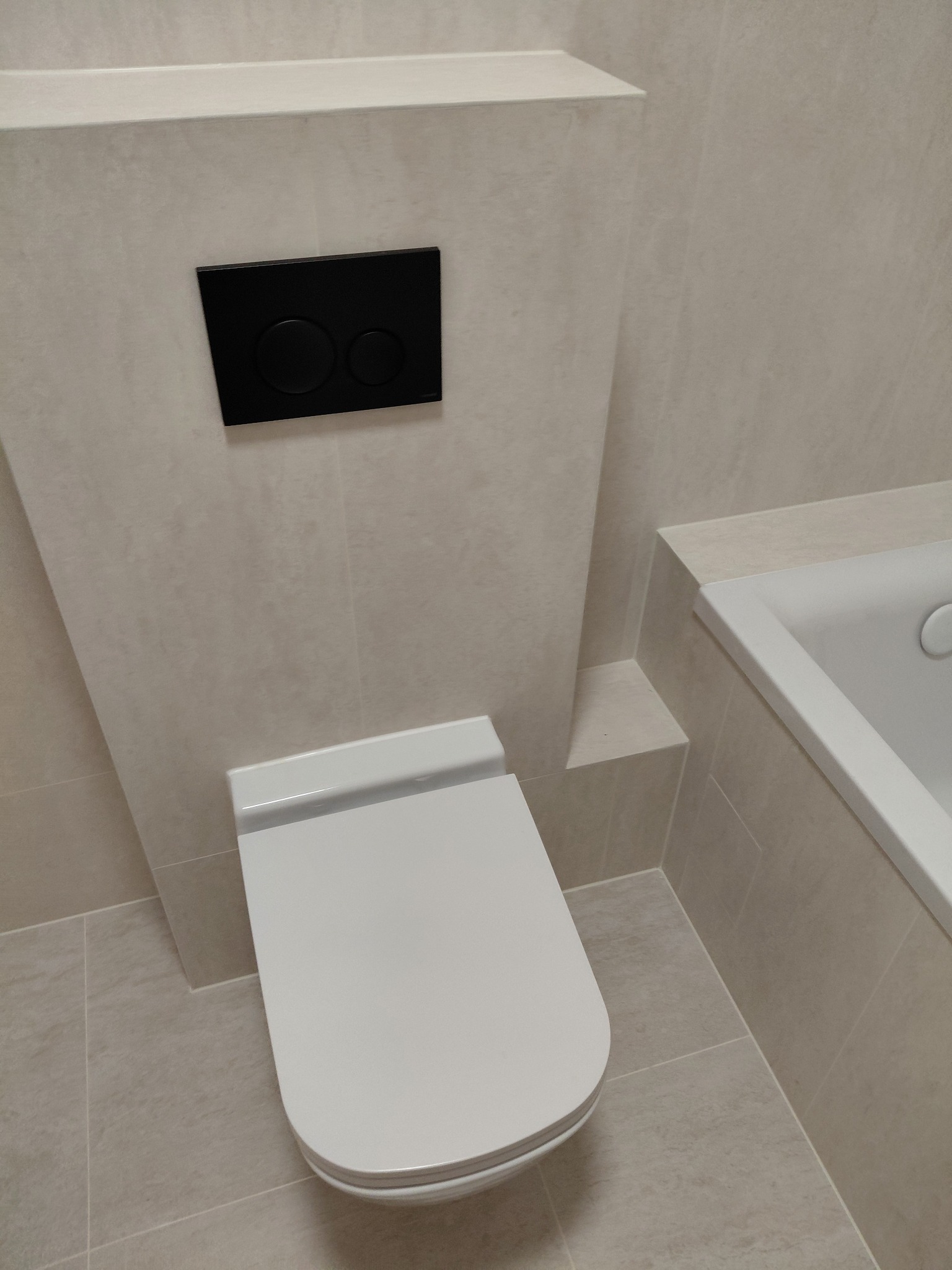 Nowoczesna toaleta podwieszana z czarnym panelem spłukującym na tle beżowych płytek w wykończonej łazience. Minimalistyczny design, czyste linie.