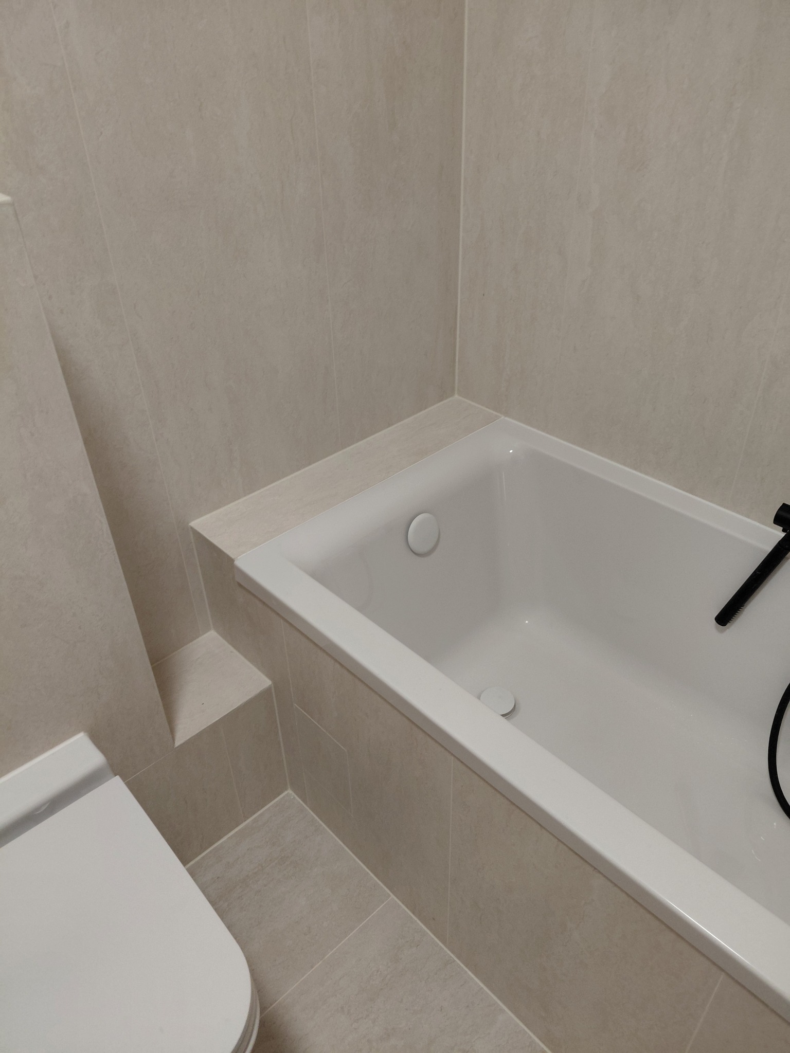 Minimalistyczna łazienka z wanną w zabudowie i podwieszaną toaletą, wykończona beżowymi płytkami imitującymi kamień. Nowoczesna armatura.