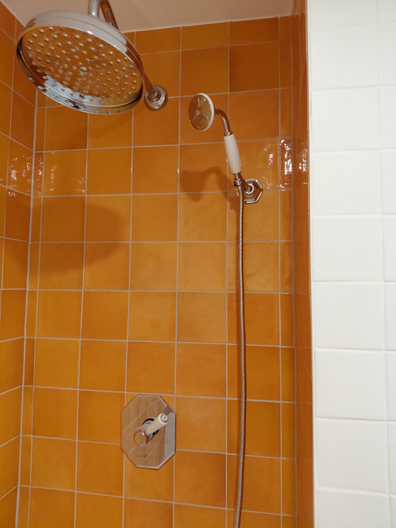 Pomarańczowe, kwadratowe płytki w kabinie prysznicowej z chromowaną armaturą i słuchawką. Część ściany wyłożona białymi płytkami. Widoczny mieszacz wody.