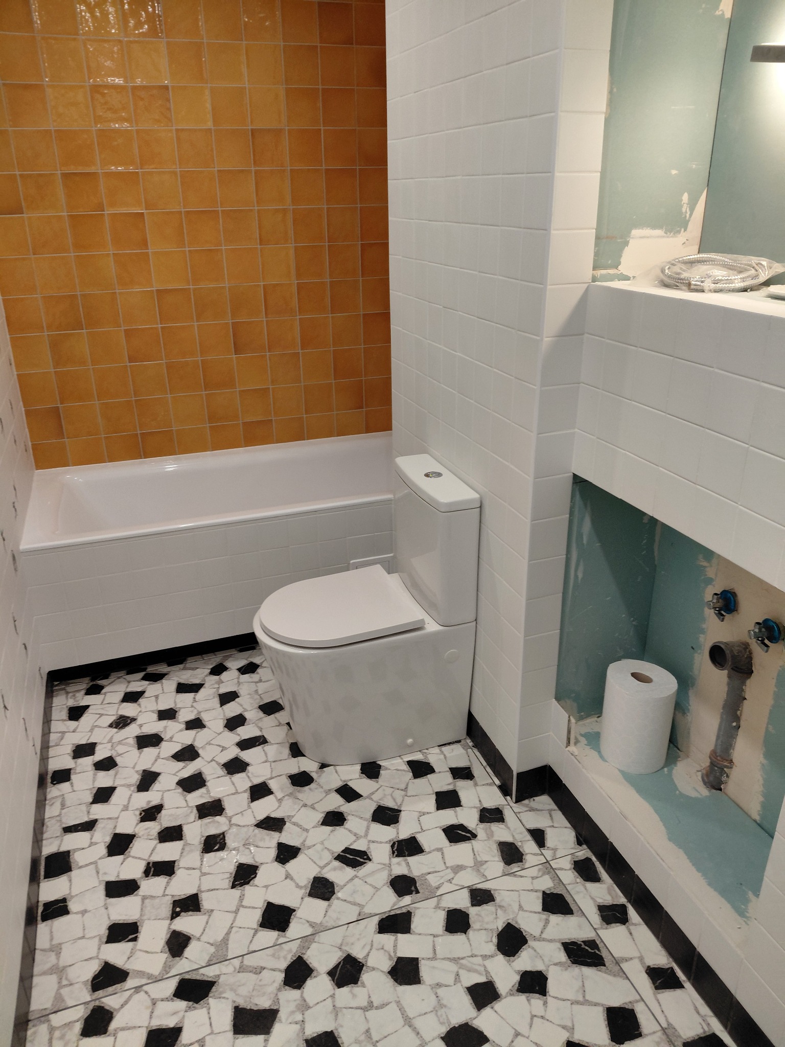 Biała toaleta w łazience z mozaikową podłogą i pomarańczowymi płytkami nad wanną. Widoczne przyłącza wodne i rolka papieru toaletowego w niszy.