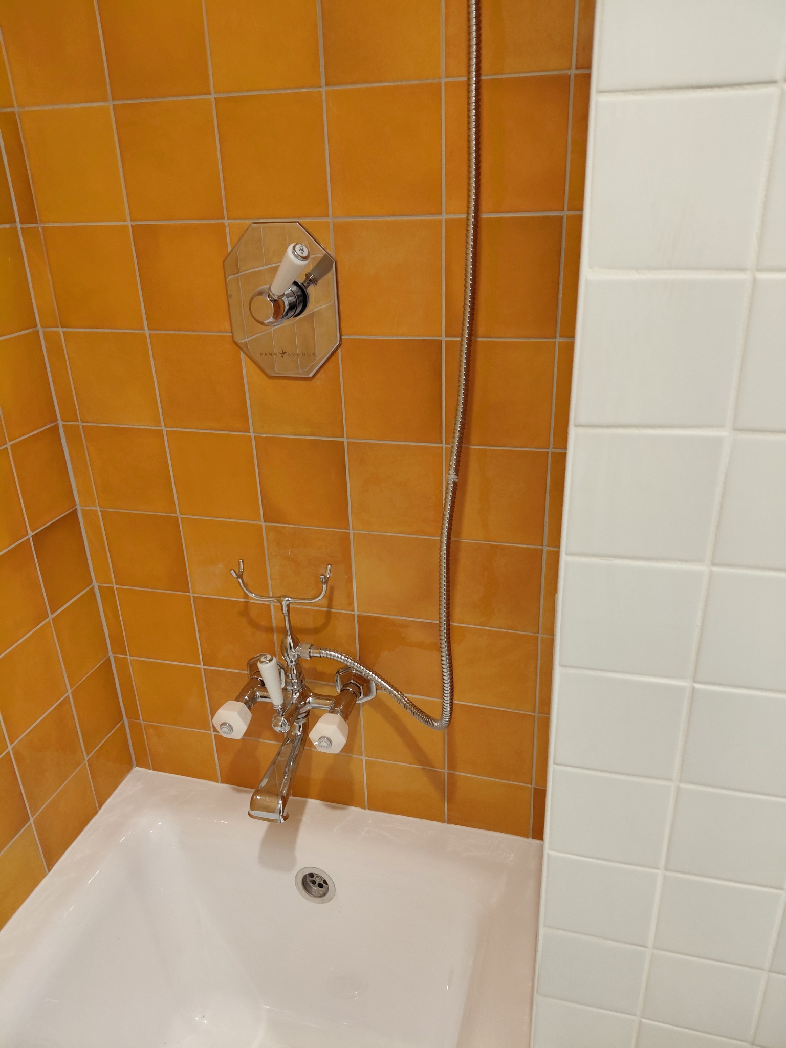 Biała wanna z chromowaną armaturą na tle pomarańczowych i białych płytek. Widoczny fragment słuchawki prysznicowej i bateria z dwoma kurkami.