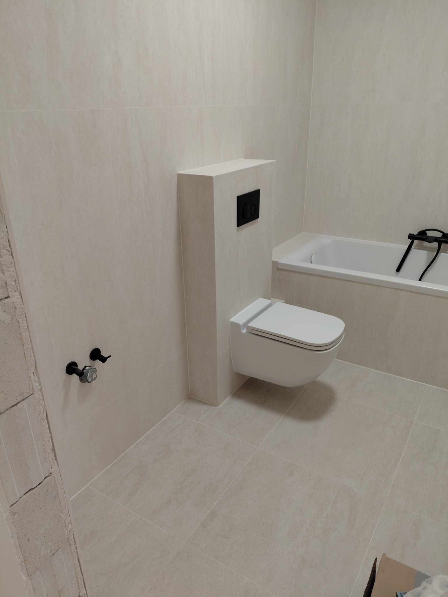 Jasna łazienka po remoncie: beżowe płytki, podwieszana toaleta z czarnym przyciskiem, bateria wannowa. Minimalistyczny design, świeżo wykończone wnętrze.