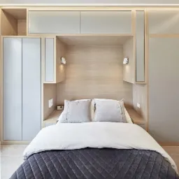 Nowoczesna sypialnia z zabudową meblową z jasnego drewna, wnęka nad łóżkiem z oświetleniem punktowym, szara pikowana narzuta i poduszki.